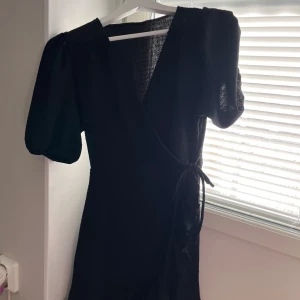Svart omlottklänning Gina Tricot M - Säljer en svart kort omlottklänning från Gina Tricot i storlek M. Klänningen har puffiga korta ärmar, v-ringning och knyt i midjan. Materialet är strukturerat och har volangdetaljer längs nederkanten för en extra snygg touch.