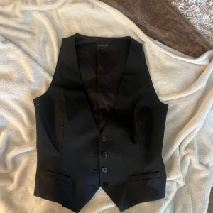 Svart kostymväst från Stockh LM - Snygg svart kostymväst från Stockh LM i klassisk modell med djup v-ringning och fem knappar framtill. Västen har två fickor fram och justerbart band med metallspänne bak. Perfekt att styla över en t-shirt eller skjorta för en cool look.