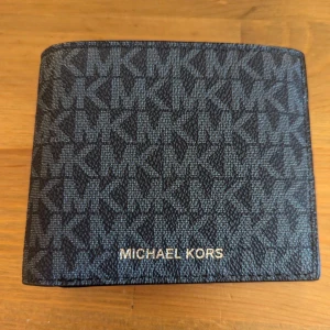 Mörkblå Michael Kors plånbok - Snygg plånbok från Michael Kors i mörkblått med MK-logga över hela ytan. Plånboken är kompakt och har en stilren design med Michael Kors i silvertext på framsidan. Materialet är slitstark syntet och mönstret ger en lyxig vibe. Nästan helt ny använd några gånger så fortfarande ganska fräsch. Pris kan diskuteras eller bud