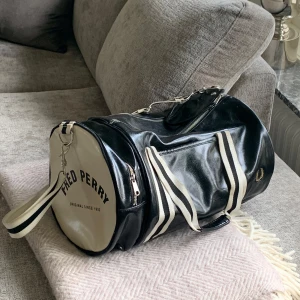 Fred Perry svart och vit axelväska - Snygg rymlig axelväska från Fred Perry i svart och vitt med rund form. Väskan har breda randiga axelremmar, dragkedja och logga på sidan. Materialet är glansigt skinn och detaljerna är i guld. Perfekt för gymmet eller när du vill ha med dig mycket. Har små fläckar på banden (se sista bild) övrigt i mycket gott skick.