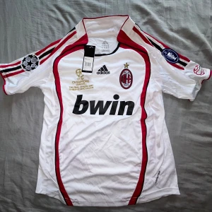 AC Milan Kaka Champions League 2007 tröja - AC Milan fotbollströja från Champions League-finalen 2007 med Kaka och nummer 22 på ryggen. Vit med röda och svarta detaljer, broderat klubbmärke och guldtext. Adidas ClimaCool-material, kortärmad och med snygga tryck på ärmarna.