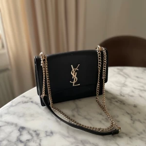 Svart YSL axelväska i skinn - Snygg svart axelväska från YSL med guldfärgad kedja och ikonisk YSL-logga framtill. Väskan är tillverkad i slätt skinn och har ett stilrent, rektangulärt format med flera fack. Perfekt för dig som gillar lyxiga detaljer och klassisk design.