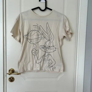 Beige Bugs Bunny t-shirt från H&M - Snygg beige t-shirt från H&M med stort tryck av Bugs Bunny från Tune Squad på framsidan. T-shirten har rund halsringning och korta ärmar, perfekt för dig som gillar Looney Tunes och vill ha en chill och lekfull stil.