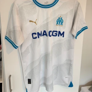 Marseille hemmatröja Puma M - Olympique de Marseille hemmatröja från Puma i vitt med ljusgrått mönster och blå detaljer. Broderat klubbmärke och guld Puma-logga på bröstet. Tillverkad i ventilerande polyester med blå/vita ribbade muddar och krage. Snygg och sportig look för alla fotbollsfans.