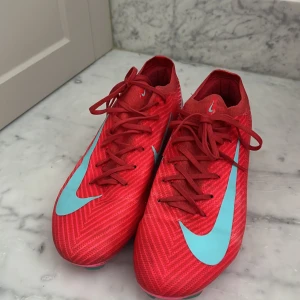 Nike Mercurial röda fotbollsskor strl 41 - Säljer ett par Nike Mercurial fotbollsskor i röd färg med turkosa detaljer och logga. Skorna har snörning, mönstrad ovandel och platt sula med gröna dobbar för gräsplan och konstgräs . Ovandelen är tillverkad i syntetmaterial och har en slimmad form som sitter tajt på foten. storleken är 41 och 26 cm, jätte bra nytt skick använt i ungefär en månad om ni vill ha nå mer detaljer Äre bara skriva:)