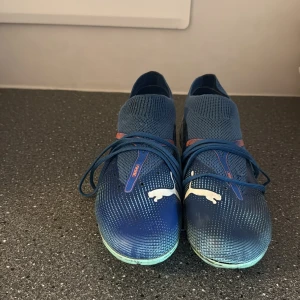 Blåa Puma fotbollsskor med snörning - Säljer ett par blåa Puma fotbollsskor med vita detaljer och logga på sidan. Skorna har snörning, stickad ovandel och mönstrad sula för bra grepp på planen. Snygg sportig design med inslag av rött och ljusblå sula. Perfekta för dig som vill sticka ut på fotbollsplanen.