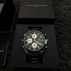 Svart kronografklocka Tommy Hilfiger - Stilren svart kronografklocka från Tommy Hilfiger med rund urtavla, silverfärgade detaljer och tre mindre vita subdials. Klockan har svart länkarmband i metall och tydliga siffror. Perfekt accessoar för dig som gillar klassisk och sportig stil.