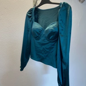 Turkos satin korsettopp med puffärm - Snygg turkos korsettopp i satin med långa puffärmar och markerade kupor. Toppen har en croppad passform och en lätt glansig yta som ger en lyxig känsla. Perfekt för dig som vill sticka ut med en trendig och elegant look.