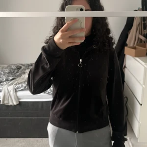 Svart velour hoodie med dragkedja - Svart hoodie i mjuk velour med huva och silverfärgad dragkedja med ringdetalj. Tyvärr har hoodien inga snören. Hoodien har långa ärmar, känguruficka framtill och ribbade muddar. Perfekt för chill dagar och snygg att styla med jeans eller mjukisbyxor.