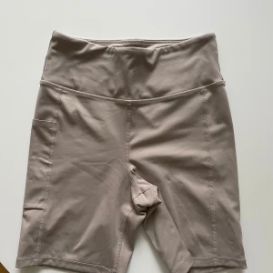 Beige träningstights-shorts Move by Gina - Snygga beige cykelshorts från Move by Gina med hög midja och praktisk ficka på sidan. Stretchigt och mjukt material som passar perfekt för träning eller chill. Söm-detaljer längs benen ger en sportig look. Perfekta för dig som gillar stilrena träningskläder.