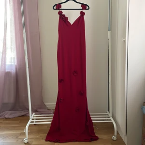 Röd långklänning med rosdetaljer - Snygg röd långklänning med v-ringning och smala axelband dekorerade med tygrosor från Nelly! Klänningen har flera rosdetaljer längs kjolen och ett elegant, flytande fall. Materialet är mjukt och har ett vackert draperat utseende 💕