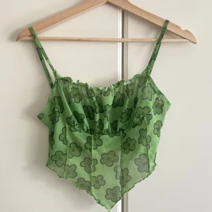 Grön blommig mesh linnetopp - Superfin grön linnetopp i mesh med blommigt mönster och smala axelband. Toppen har en spetsig nederkant och synliga sömmar framtill för en trendig look. Perfekt för dig som vill sticka ut med en färgglad och lekfull stil.