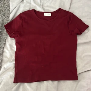 Vinröd t-shirt från Gina Tricot - En enkel och snygg vinröd t-shirt från Gina Tricot, Basically Basics-kollektionen. Modellen är klassisk med rund hals och korta ärmar. T-shirten har en normal passform och är gjord i mjuk bomull som känns skön mot huden. Perfekt till jeans eller kjol.