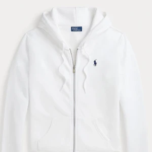 Vit hoodie från Polo Ralph Lauren - Klassisk vit hoodie från Polo Ralph Lauren med dragkedja framtill, huva med snörning och känguruficka. Broderad marinblå logga på bröstet. Perfekt för en clean och stilren look.