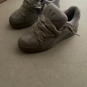 Grå chunky sneakers med bred snörning - Säljer ett par grå chunky sneakers med breda snören och coola detaljer. Skorna har en robust sula och är tillverkade i en mix av syntet och textil. Perfekta för dig som gillar streetstyle och vill ha en clean look. De är Bershka 