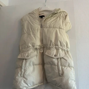 Beige dunväst med huva från New Look - Snygg beige dunväst från New Look med huva och dragkedja framtill. Västen har två stora fickor med lock och snörning i midjan för en justerbar passform. Perfekt att slänga över en hoodie för en cool och avslappnad look.