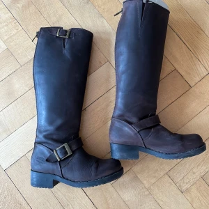 Johnny Bulls - Bruna höga läderstövlar - Snygga höga stövlar i mörkbrunt skinn med grov sula och låg klack. Stövlarna har dekorativa spännen i mässing och dragkedja baktill för enkel påtagning. Mycket sparsamt använda 