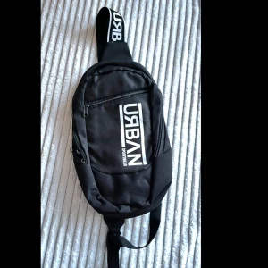 Svart Urban axelväska med tryck - Snygg svart axelväska från Urban Sportswear med vit logotyp på framsidan och bred axelrem med Urban-tryck. Väskan har flera fack med dragkedja och är gjord i slitstark polyester. Perfekt för att bära med sig det viktigaste.