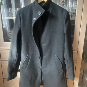 Svart rock från Bruun & Stengade - Svart rock från Bruun & Stengade i slim fit. Rocken har en hög krage med två knappar, snedställda fickor och stängs med knappar framtill. Tillverkad i en blandning av ull och polyester vilket gör den både stilren, varm och hållbar.  Rocken är knappt använd och i mycket fint skick. En avtagbar innerväst ingår (ej med på bild). Nypris ca 3000 SEK.