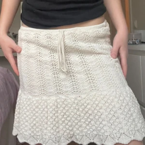 Vit stickad kjol med volangkant - Supersöt vit stickad kjol med volangkant och snörning i midjan. Kjolen har ett fint virkat mönster och känns riktigt somrig och chill. Perfekt till en enkel topp eller hoodie för en avslappnad look.