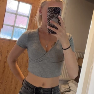Grå croppad omlottopp - Säljer en ljusgrå croppad topp med omlott-skärning och v-ringning. Toppen är ribbad, har korta ärmar och sitter tight. Perfekt för dig som gillar en enkel men trendig stil.