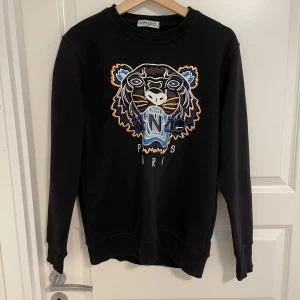 Svart Kenzo sweatshirt med tigerbrodyr - Svart sweatshirt från Kenzo med ikonisk tigerbrodyr i blått, orange och vitt framtill. Tröjan har rund hals, långa ärmar och ribbade muddar. Perfekt statement-plagg för dig som vill sticka ut med cool design och snygga detaljer.