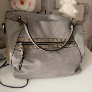 Grå axelväska med nitar och dragkedja - Snygg grå axelväska i syntetmaterial med guldfärgade nitar och detaljer. Väskan har en dekorativ dragkedja framtill och en praktisk ficka med dragkedja på baksidan. Perfekt storlek för att få plats med allt du behöver och cool design med svarta kanter.