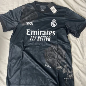 Real Madrid x Y-3 svart fotbollströja - Snygg svart Real Madrid fotbollströja från Adidas Y-3 kollektionen, designad av Yohji Yamamoto. Tröjan har korta ärmar, blommigt mönster med rosor och klassiska loggor på bröstet. Tillverkad i lätt och ventilerande material med Aeroready-teknologi.