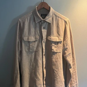 Beige Overshirt från Riley - Säljer en stilren beige overshirt från Riley i linne. Skjortan har klassisk krage, två stora bröstfickor med lock och knäppning framtill. Perfekt att slänga på sig over tjocktröja eller tshirt. Långärmad och i normal passform. Kom privat för frågor och bilder.