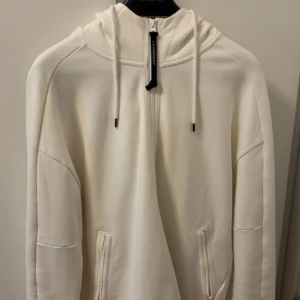 Vit hoodie från C.P. Company med goggles - Säljer en clean vit hoodie från C.P. Company med ikoniska goggles på huvan. Tröjan har half zip, dragsko i huvan och två fickor med dragkedja. Perfekt för dig som vill sticka ut med streetwear-vibe. Materialet känns mjukt och skönt, troligen bomull. Storlek M eller L minns inte riktigt.