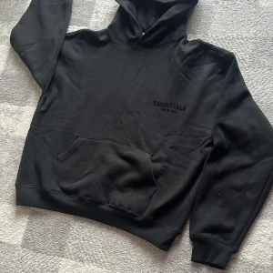 Svart Essentials Fear of God hoodie - Svart hoodie från Essentials Fear of God med broderad logga på bröstet och ryggen. Klassisk känguruficka framtill, huva och ribbade muddar. Perfekt för en clean och stilren streetwear-look.