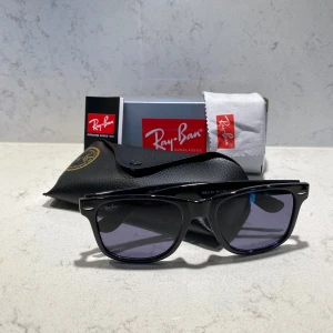 Svarta Ray-Ban Wayfarer solglasögon - Ray-Ban Wayfarer RB2140 solglasögon med klassisk svart båge i plast och mörka glas. Ikonisk design med silverdetaljer vid fronten. Levereras med originalfodral och putsduk. Perfekt för dig som vill ha en tidlös och cool look.