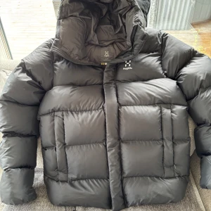 Svart dunjacka från Haglöfs - Säljer en svart, riktigt fluffig dunjacka från Haglöfs med stor huva och diskret logga på bröstet. Jackan har breda, quiltade paneler och dragkedja framtill. Perfekt för kalla dagar när du vill hålla värmen och ändå se clean ut.