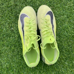 Nike Air Zoom fotbollsskor neongula - Snygga neongula Nike Air Zoom vapor elite. fotbollsskor med svart swoosh och vita detaljer. Ovandelen är i syntetmaterial med snörning och mönstrad textur för extra grepp. Sulan har fasta dobbar och transparent finish, perfekt för spel på gräs. Lätta och smidiga för snabba rörelser på planen.