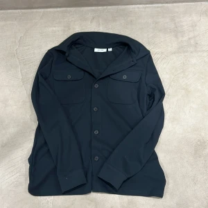 Svart overshirt från Only & Sons - Stilren svart overshirt från Only & Sons med klassisk krage, två bröstfickor med lock och knappar framtill. Skjortan har lång ärm och är tillverkad i ett mjukt bomullstyg. Perfekt att slänga över en t-shirt för en avslappnad look.