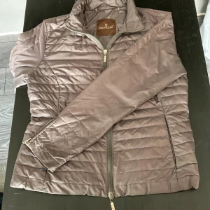 Grå quiltad pufferjacka från Moncler - Snygg grå pufferjacka från Moncler med quiltad design och dragkedja framtill. Jackan har hög krage, två sidofickor med dragkedja och är lätt vadderad. Perfekt för kyliga dagar när du vill ha både stil och komfort. (Gammal modell, säljs ej längre, 100% ÄKTA)