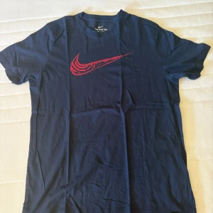 Mörkblå Nike t-shirt med röd logo - Snygg mörkblå t-shirt från Nike med klassisk röd swoosh på bröstet. Modellen är kortärmad och har en rund hals. Perfekt för dig som gillar sportiga och stilrena plagg med tydlig logga. Materialet känns mjukt och skönt mot huden.