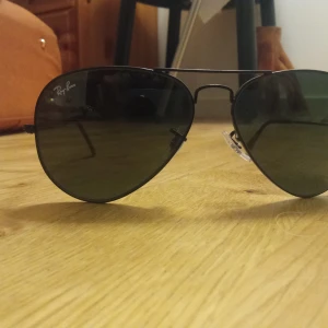 Svarta Ray-Ban aviator solglasögon - Snygga svarta Ray-Ban aviator solglasögon med klassisk pilotform. Tunn metallbåge och mörka linser som ger en cool vibe. Passar perfekt för dig som vill ha en tidlös och stilren look. Ikonisk Ray-Ban-logga på linsen.