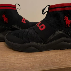 Svarta Polo Ralph Lauren sneakers - Svarta sneakers från Polo Ralph Lauren med chunky sula och elastiskt band över foten med röd POLO-logga. Stickad ovandel med röd broderad logga på sidan och ribbad mudd i svart och rött. Snygg sportig look med detaljer i skinn och syntet. Köpt på NK Stockholm för 2490kr är i nyskick! 