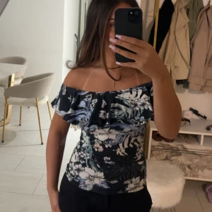 Blommig offshoulder topp Gina Tricot - Supersöt offshoulder topp från Gina Tricot med blommigt mönster i svart, vitt och blått. Toppen har volang upptill och sitter tight över kroppen. Perfekt för dig som gillar en feminin och trendig stil.