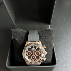 Seiko daytona mod - Snygg Seiko kronograf med svart urtavla och guldiga detaljer. Klockan har tre subdials, svarta och guldiga visare samt index med stenar. Boetten är i guldton och armbandet är svart och gjort i mjukt material. Perfekt accessoar för dig som gillar sportig och lyxig stil.