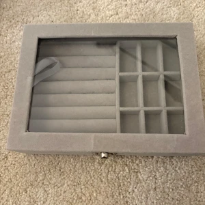 Grå smyckesbox med glaslock - Snygg och stilren smyckesbox i ljusgrå sammet med glaslock. Perfekt för att organisera ringar, örhängen och andra småsmycken. Boxen har flera fack och mjuka rader för att hålla dina favoriter på plats. Liten metallknäppning framtill.