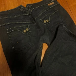 Y2k Lågmidjade bootcut jeans  - Säljer ett par svarta jeans från River Island med bootcut passform och snygga detaljer som dubbla knappar och kontrastsömmar. Jeansen har fickor med dekorativa nitar och broderad text på insidan av linningen. Perfekta för dig som gillar en cool och trendig look.