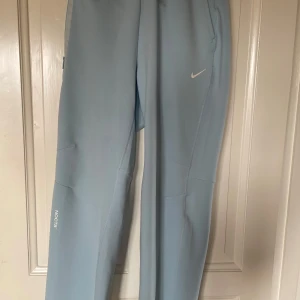 Nike NOCTA ljusblå mjukisbyxor - Säljer ett par ljusblå mjukisbyxor från Nike NOCTA med diskreta vita loggor och tryck. Byxorna har elastisk midja, raka ben och är tillverkade i ett mjukt syntetmaterial. Perfekta för chill eller träning, med stilren och clean look. Färg Baby Blue