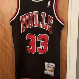 Chicago Bulls 1997-98 Jersey  - Säljer en svart Chicago Bulls baskettröja från Mitchell & Ness Hardwood Classics, med nummer 33 och Pippen på ryggen. Tröjan har röda och vita detaljer samt stora tryck fram och bak. Perfekt för dig som älskar basket och vill ha en ikonisk look.