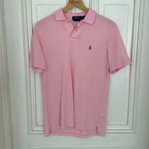 Rosa pikétröja från Polo Ralph Lauren - Snygg rosa pikétröja från Polo Ralph Lauren med klassisk krage och två knappar. Tröjan har korta ärmar och det ikoniska blå broderade logot på bröstet. Tillverkad i mjuk bomull som är skön mot huden. Perfekt för en chill och stilren look.