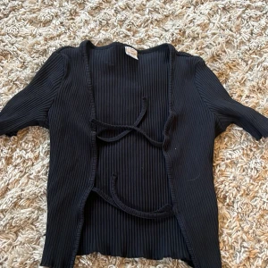 Svart ribbad kofta från Lindex - Svart ribbad kofta från Lindex med knytband framtill. Toppen har korta ärmar och är tillverkad i ett mjukt, stretchigt material. Man kan ha den öppen men även knyt. 