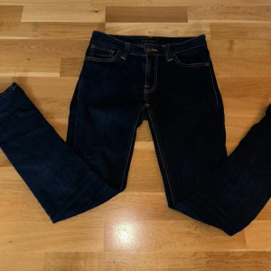 Mörkblå jeans från Nudie Jeans, 27/30 - Säljer ett par mörkblå jeans från Nudie Jeans med klassiska orange sömmar och snygga bakfickor med vågig brodyr. Modellen har normal passform och raka ben. Tillverkade i slitstark bomull med en stilren look som funkar till det mesta.