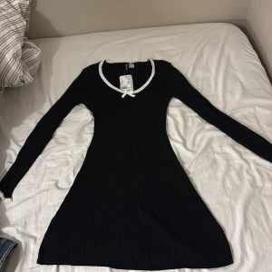 Svart ribbad kortklänning med rosett - Supersöt svart ribbad kortklänning från H&M med lång ärm och v-ringning. Kragen är dekorerad med vit spetskant och en liten rosett framtill. Klänningen har en figurnära passform och är perfekt för dig som gillar en enkel men gullig stil.