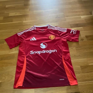 Manchester United Adidas fotbollströja röd - Snygg Manchester United fotbollströja från Adidas i rött med vita detaljer och klubbmärke på bröstet. Tröjan har korta ärmar, rund hals och är tillverkad i lätt och ventilerande material. Stora loggor på bröstet och ärmen samt vita ränder på axlarna.